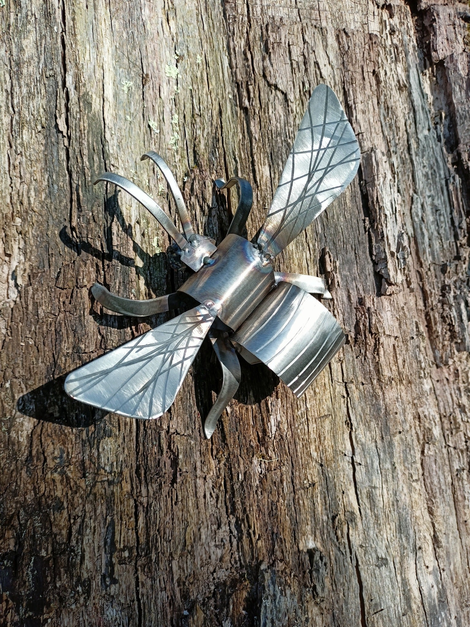 A lacquered stainless steel bee artwork placed outdoors, clinging to a tree stump