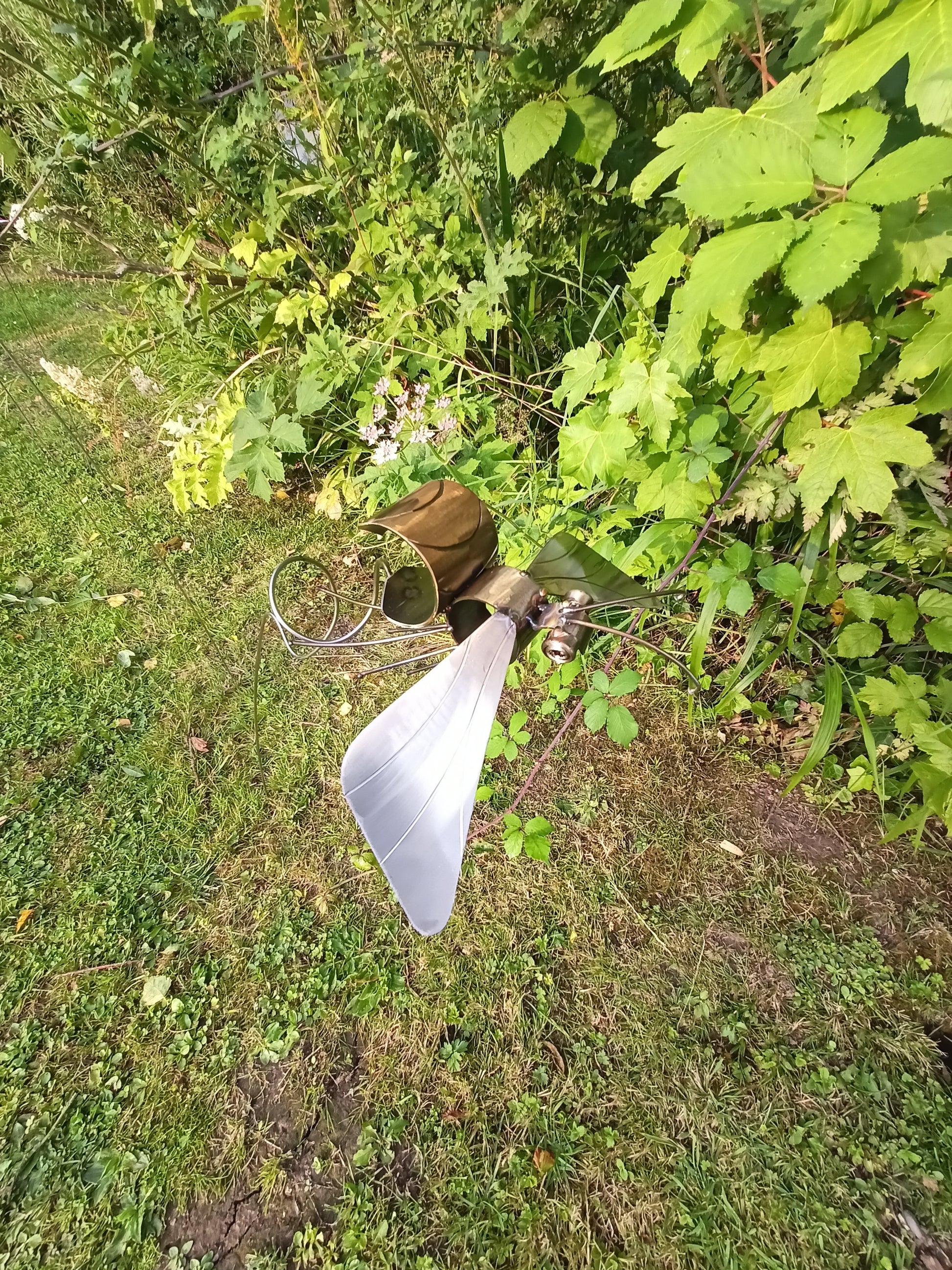 A lacquered stainless steel bee artwork placed outdoors, surrounded by greenery and in mid flight.
