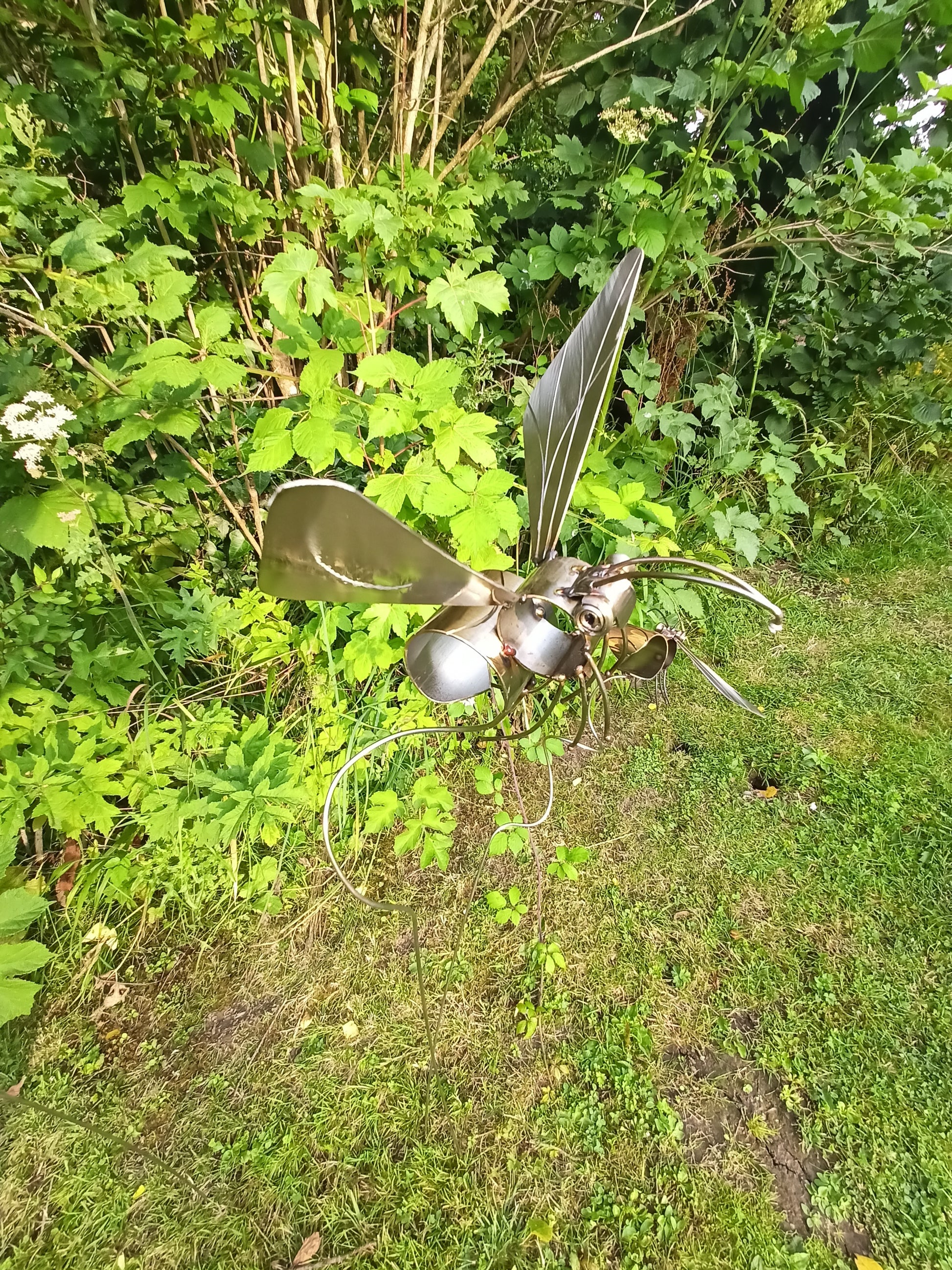 A lacquered stainless steel bee artwork placed outdoors, surrounded by greenery and in mid flight.