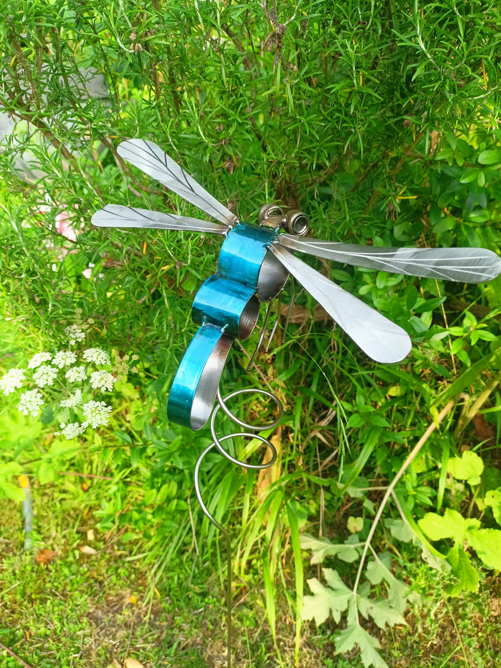 A metal dragonfly sculpture with blue and silver colors, positioned outdoors among greenery.