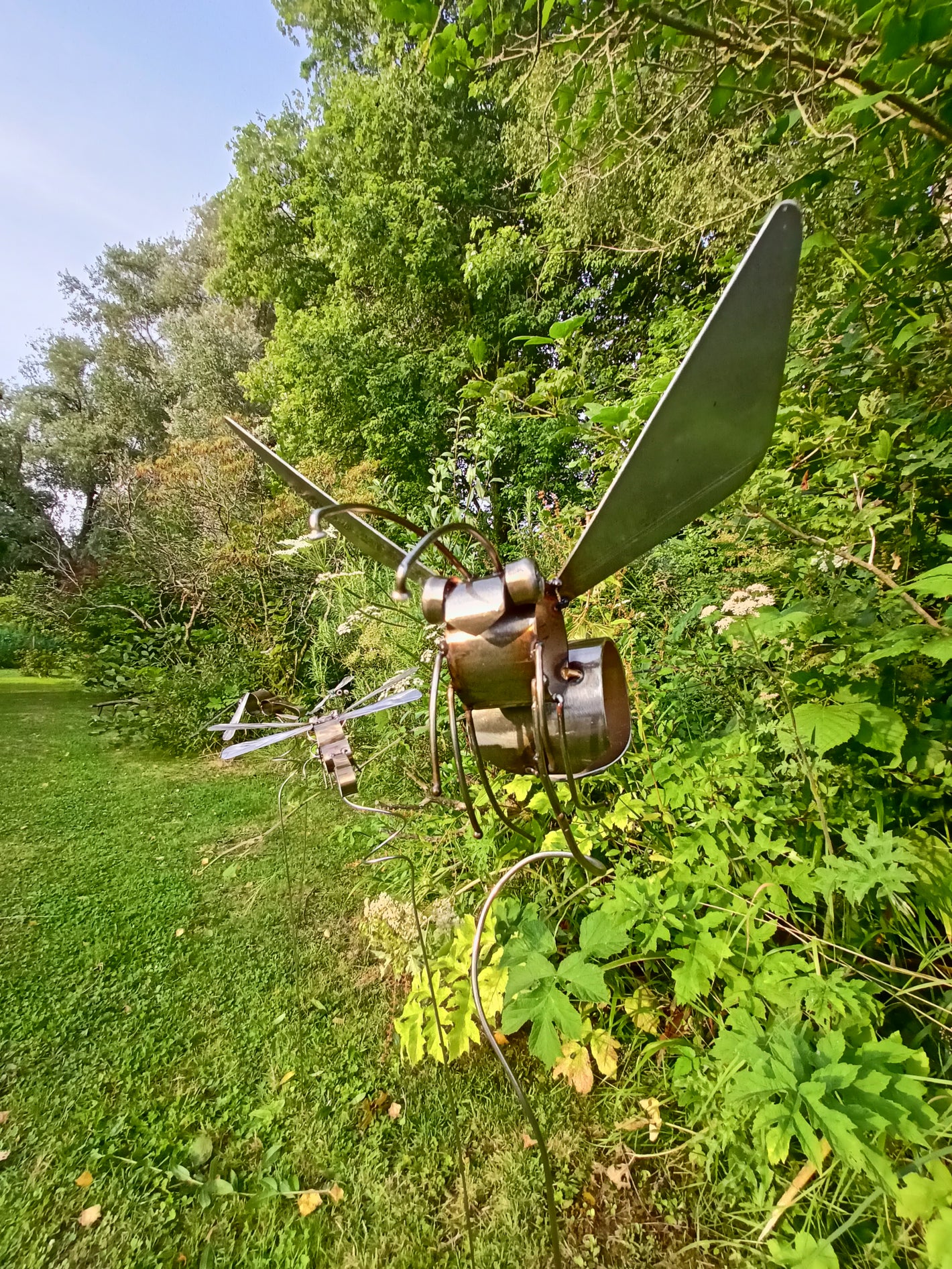 A lacquered stainless steel bee artwork placed outdoors, surrounded by greenery and in mid flight.